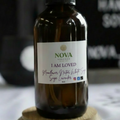 Bottle of Nova product labeled 'I Am Loved' on a wooden stand with a blurred background.