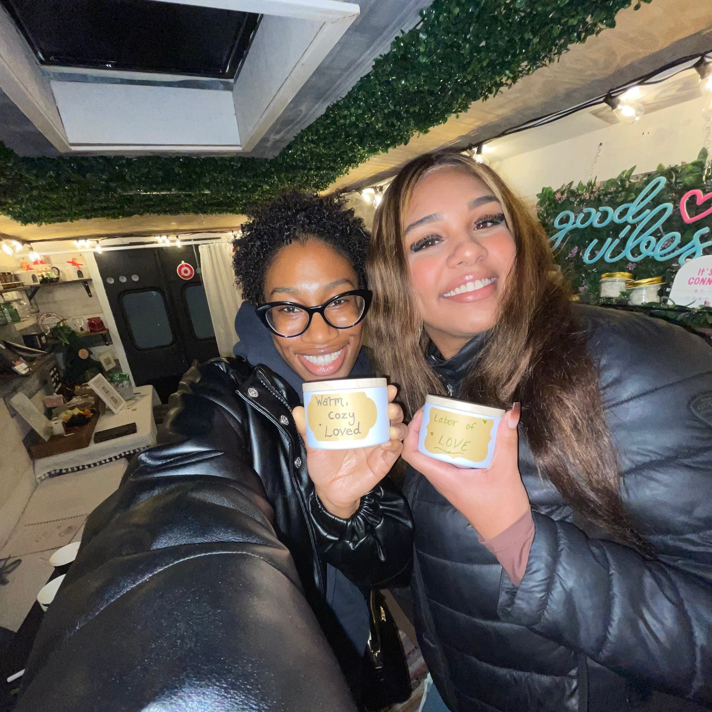 Two women holding small containers with a 'good vibes' sign in the background.