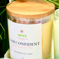 Candle with wooden lid labeled 'I am Confident' by Nova Essence on a white surface with greenery in the background.