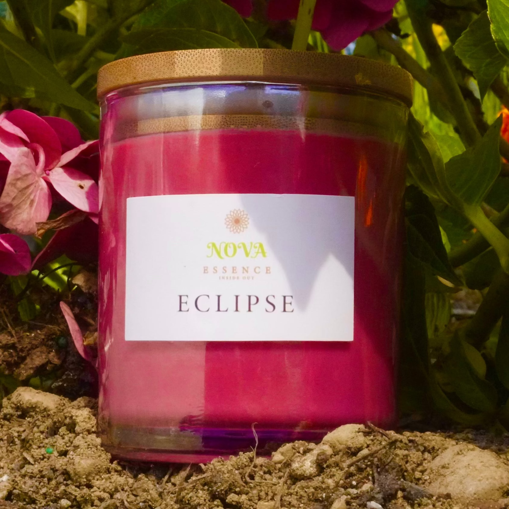 Candle labeled 'Eclipse' by Nova Scented in a garden setting with pink flowers.